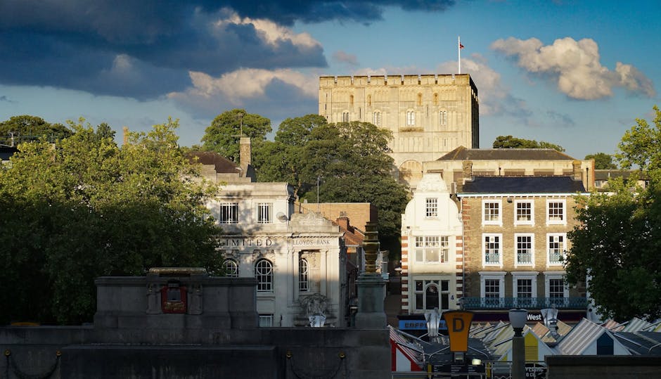 A cityscape scene in Kingston upon Thames featuring historic buildings and a castle tower in the background. In the foreground, there are trees with lush green foliage and a stone wall with an ornate lantern. The midground displays a row of commercial and residential buildings with varying architectural styles, including brick and stucco facades, some with decorative window frames. The sky above shows a mix of blue and dark clouds, indicating an overcast weather. This setting illustrates a typical urban environment where house removals and furniture transport may take place, with a focus on the local architecture and street scene relevant to services such as packing and moving, as provided by removal companies like [COMPANY_NAME].
