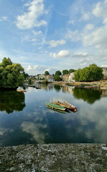 A cityscape scene in Kingston upon Thames featuring historic buildings and a castle tower in the background. In the foreground, there are trees with lush green foliage and a stone wall with an ornate lantern. The midground displays a row of commercial and residential buildings with varying architectural styles, including brick and stucco facades, some with decorative window frames. The sky above shows a mix of blue and dark clouds, indicating an overcast weather. This setting illustrates a typical urban environment where house removals and furniture transport may take place, with a focus on the local architecture and street scene relevant to services such as packing and moving, as provided by removal companies like [COMPANY_NAME].
