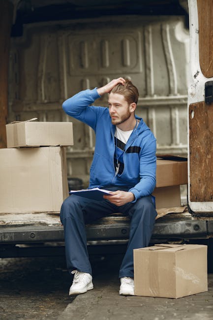 A young man with light brown hair and a beard, wearing a blue hoodie, dark blue trousers, and white sneakers, is sitting on the edge of an open moving van. He is holding a clipboard in his left hand and appears to be contemplating or making notes, with his right hand resting on his head. Surrounding him are several cardboard boxes, some stacked inside the van and others placed on the pavement outside. The interior of the van is visible in the background, showing a spacious area with a beige panel and a metal floor. The scene is set outdoors during daylight, with natural lighting highlighting the packing and home relocation process managed by a professional removals service like [COMPANY_NAME]. This image depicts the logistical planning and packing stage involved in house removals, emphasizing careful handling and organization of belongings during a furniture transport or moving operation.