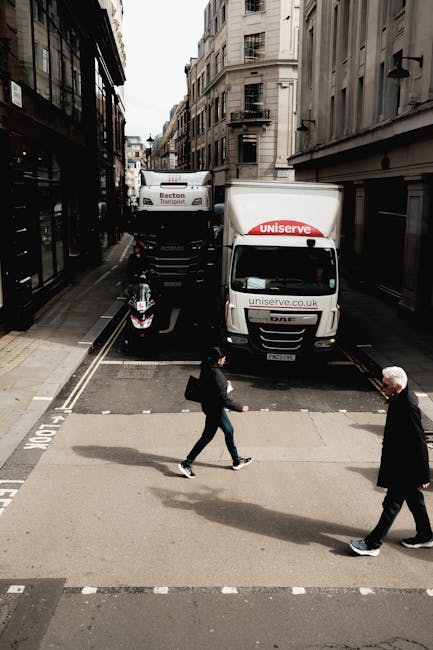 A city street scene showing a narrow, one-way road lined with tall buildings on both sides, with a small, designated loading area at the curb. In this area, a white moving van branded with 'Uniserve' and 'uniserve.co.uk' on its side is parked with its rear doors open, likely during a home relocation or furniture transport process. A black motorcycle is parked nearby, close to the van. Two pedestrians are crossing the street at a pedestrian crosswalk; one is a woman dressed in a dark jacket and jeans carrying a bag, and the other is an older man with a white beard, wearing a black jacket and dark trousers. The scene appears to be during daytime with natural lighting, capturing the typical logistics involved in professional removals, including vehicle loading and pedestrian movement, reflecting aspects of packing and moving operations carried out by Kingston-based removal companies.