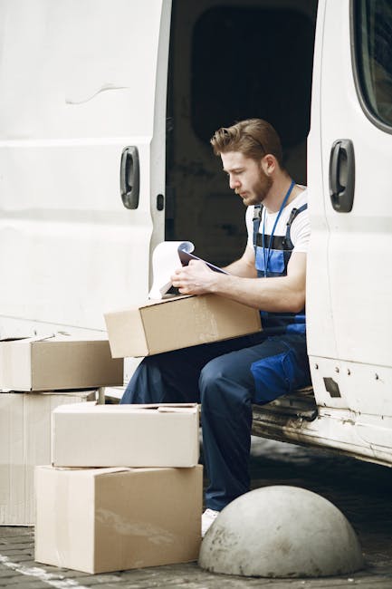 A man dressed in casual work clothing, sitting on the edge of an open white delivery van with the side door open, surrounded by several opened cardboard moving boxes on the pavement. He is holding a c