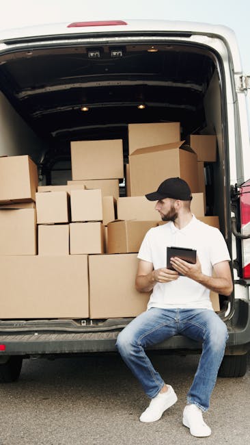 A man dressed in casual work clothing, sitting on the edge of an open white delivery van with the side door open, surrounded by several opened cardboard moving boxes on the pavement. He is holding a c