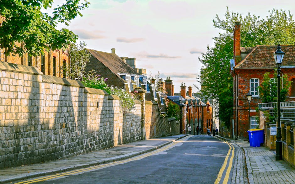 A narrow residential street during daytime featuring a stone wall on the left side with lush green trees and flowering plants extending over the top. On the right side, there are traditional brick houses with chimneys, some with pitched roofs, and a black street lamp nearby. The road has double yellow lines along the curb, indicating parking restrictions, with a blue recycling bin positioned by the pavement. The street surface is asphalt, with a concrete sidewalk on the right side. In the distance, a small group of people is visible, along with parked vehicles and a van partially visible at the far end. The scene depicts a quiet, typical UK street, suitable for home relocation activities involving furniture transport and loading processes, aligning with [PAGE_TITLE] from [DOMAIN]. The lighting suggests late afternoon with soft sunlight casting shadows, emphasizing the textures of the brickwork and stone wall.