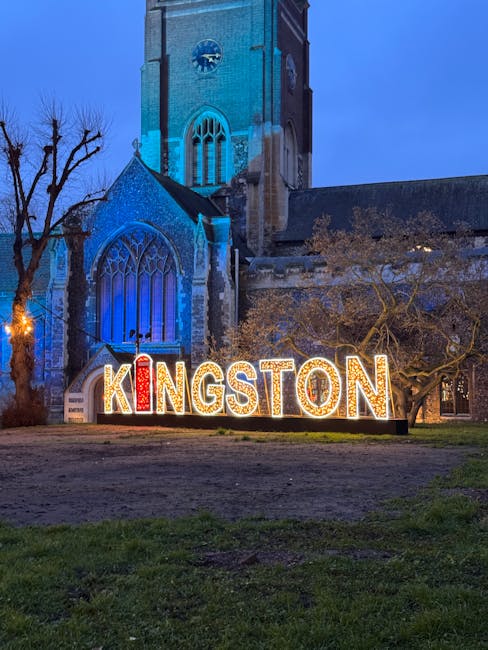 Nighttime view of Kingston signage in front of a historic church with a tall steeple and large stained glass windows, illuminated by exterior lighting and blue uplighting on the church façade. The sign features large, illuminated letters spelling 'KINGSTON' positioned on a grassy area, with a leafless tree nearby. The scene captures a quiet, well-lit street in Kingston upon Thames, suggesting a promotional display or landmark near a house removal and furniture transport service, with the church as a backdrop during evening hours.