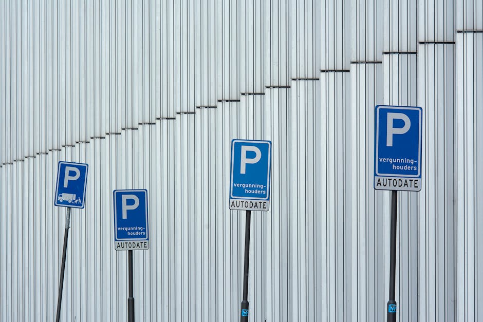 Four blue parking permit signs mounted on black metal poles are positioned in a row in front of a modern, vertically ribbed metallic wall. The signs indicate parking permissions with a prominent white