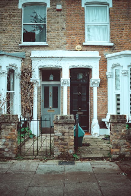 The photograph shows the front of a two-storey terrace house with a brick exterior and two large sash windows on the upper floor. The ground floor features a central porch area with a white-painted decorative architrave and columns, framing the front door, which has a glass panel and a brass letterbox. To the right of the door, there is a small step leading onto the paved pathway, and a nearby black metal fence with stone pillars separates the property from the sidewalk. A green delivery box is positioned on the pathway near the entrance. The house's facade shows some climbing plants on the left side, and the surrounding area includes neighboring terrace houses with similar architectural details. This scene depicts a typical residential setting suitable for house removals and furniture transport, emphasizing the confined front access often encountered during home relocation efforts for terrace homes in Kingston upon Thames.