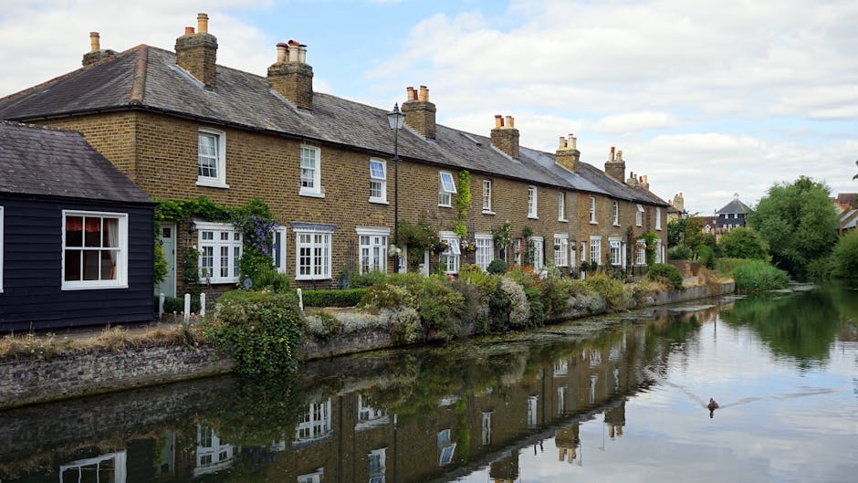 A row of traditional terraced houses constructed from yellow brick with white-framed windows and chimneys, situated along a canal or river in Kingston upon Thames. The houses have small front gardens with greenery, climbing plants, and garden ornaments, and are directly accessible from the pavement. A black wooden outbuilding is visible on the left side of the image, close to the water's edge. The water reflects the houses, sky, and surrounding trees, with a duck swimming in the foreground. The scene is captured during daytime with partly cloudy weather, showcasing a peaceful residential environment. This setting illustrates potential challenges faced during house removals or furniture transport in terrace homes, such as narrow access paths and confined delivery areas, emphasizing the importance of professional removal services like those provided by Kingston removals.