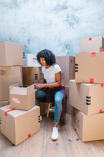 A young woman with long dark hair, sitting on a wooden floor inside a room, surrounded by numerous large cardboard moving boxes of varying sizes, some sealed with red packing tape and others with prin