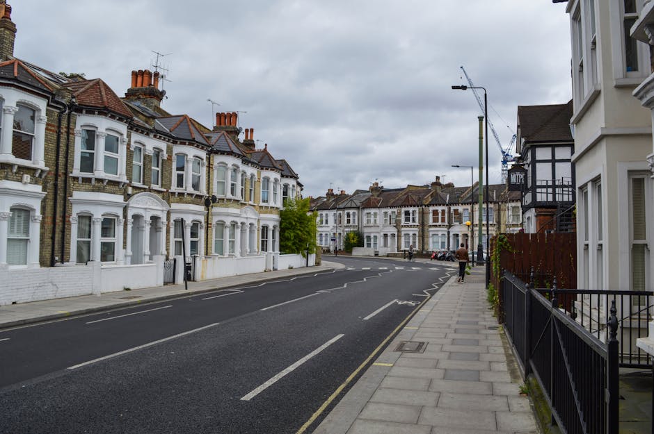 Photograph of a residential street during daytime with a row of Victorian-style terraced houses on the left, featuring white facades, bay windows, and brown tiled roofs. The scene shows a paved sidewalk with black metal railings on the right, alongside attached houses, and a wide, empty asphalt road with lane markings. Overhead, there are grey, overcast skies. In the background, a construction crane and modern street lighting posts are visible, indicating ongoing development or renovations nearby. This image captures a typical UK street setting that could relate to home relocations or house removals, emphasizing the environment where furniture transport and packing could occur before moving day, with details suitable for content about moving logistics and residential relocation services by companies like [COMPANY_NAME].