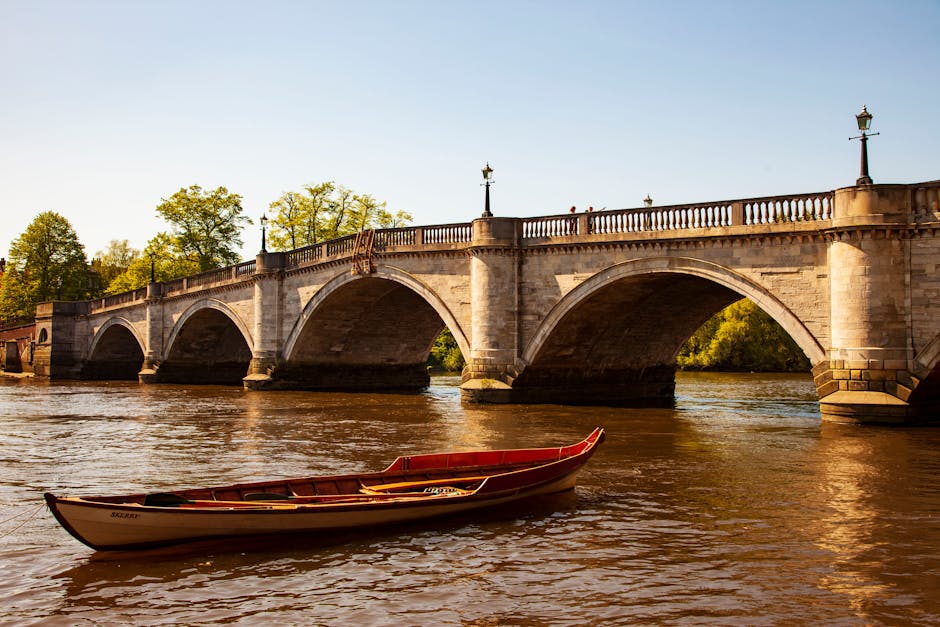 A red and white wooden rowboat floating on calm water near the base of a historic stone bridge with multiple arches, decorative lampposts along the bridge railing, and lush green trees in the background. The scene is captured in natural daylight, highlighting the textures of the stonework and the gentle ripples in the water. The setting suggests a peaceful riverside environment, which may be relevant for house removals or relocation logistics involving water transport or scenic surroundings in Kingston upon Thames, as detailed in the Kingston removals guide for Richmond Road and Canbury residents on removalcompanieskingstonuponthames.co.uk.