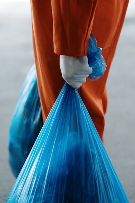 Close-up of a person wearing orange overalls and a white glove, holding a blue plastic rubbish bag filled with waste, with the background featuring a blurred outdoor setting. The individual appears to be involved in waste disposal or removal activities, possibly related to home relocation or clearing a property. The scene emphasizes careful handling of waste materials, which may be part of the packing, moving, or disposal process during house removals. This image supports the understanding of proper waste disposal practices in line with Kingston waste disposal rules for removals, as highlighted on the Kingston upon Thames moving services webpage. The focus is on the hand gripping the bag, the protective glove, and the practical aspect of managing waste efficiently outside a property, aligning with the general themes of furniture transport, packing, and loading during a house move by companies like [COMPANY_NAME].