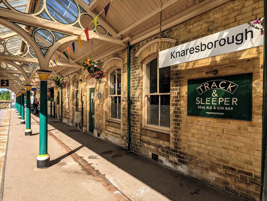Photograph of Knaresborough railway station platform in daylight, showing a row of green and cream decorative cast-iron columns supporting a detailed arched canopy with intricate patterns and hanging flower baskets. The platform surface is paved with concrete, and there are a few people sitting on benches along the edge, waiting for a train. A large white sign with black text indicates 'Knaresborough,' and underneath, a dark green sign displays 'Track & Sleeper, Real Ale & Gin Bar,' with the station's name repeated. The brick exterior of the station building features arched sash windows with white frames, and the overall scene conveys a calm, organized environment suitable for a home relocation or moving service context involving transportation and logistical planning from a traditional station setting.