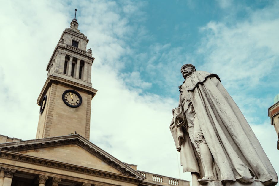 A historic stone clock tower with a pointed spire and a large round clock face displaying the time as 4:35 is visible in the background, set against a partly cloudy sky. In the foreground, a classical-style stone statue of a man dressed in period attire, including a long cloak and holding a satchel, is positioned on a pedestal. The scene depicts part of a historic building with a triangular pediment supported by columns. This image naturally relates to house removals and relocation services in Kingston upon Thames, illustrating the area’s architectural heritage, and can be associated with the logistics of furniture transport and home relocation processes that companies like [COMPANY_NAME] may handle in the vicinity of Kingston station, as referenced on the page titled 'KT1 flat removals advice near Kingston station, KINGSTON UPON THAMES'.