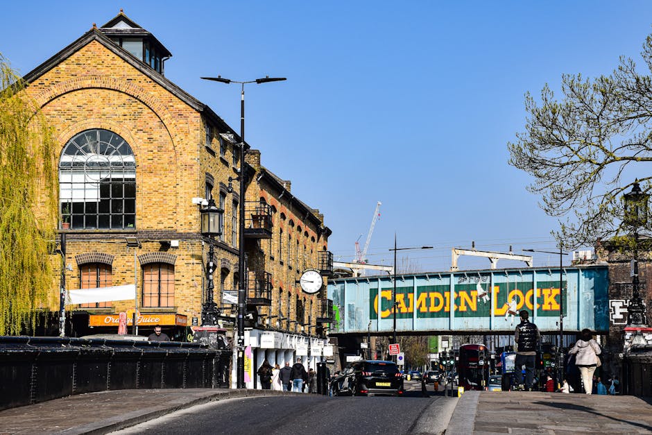 A street scene in Kingston upon Thames showing a large brick building with a prominent arched window and a clock mounted on the wall. Several pedestrians are walking along the sidewalk, some carrying bags, while others are standing near storefronts such as a juice shop. The street features parked cars, and a blue metal footbridge with the words 'Caladin Lock' painted in yellow spans across the road above a road closure barrier. Streetlights and leafless trees are visible, with the sky clear and blue overhead. This setting illustrates an urban environment suitable for house removals, with a focus on loading and unloading activities typical of home relocation and furniture transport, connected to quick, local moving services like those provided by [COMPANY_NAME] in Kingston upon Thames.