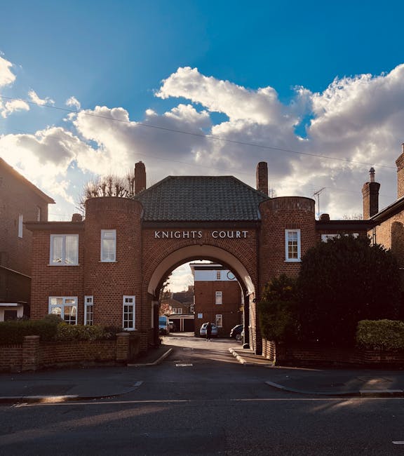 A brick-built residential building with an arched entrance labeled 'Knights Court' serving as a gateway to a courtyard area. The structure features multiple windows, some with white frames, and a pitched roof with chimney stacks. In the foreground, a paved driveway leads through the arch, with parked cars visible inside the courtyard. The scene is set against a partly cloudy sky with sunlight illuminating the building, and greenery such as shrubs on either side of the entrance. This setting represents a typical home relocation or apartment complex often involved in house removals or furniture transport, with the imagery underscoring the process of loading or unloading within a secure residential environment coordinated by a professional removal service like [COMPANY_NAME].