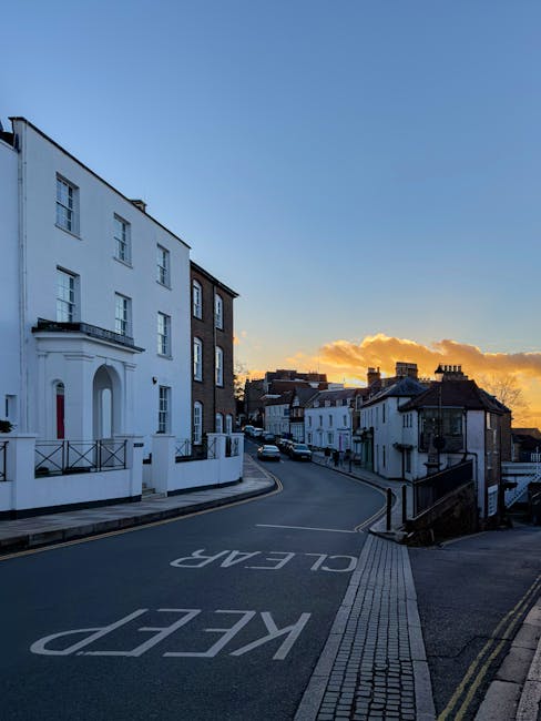 A quiet residential street in Surbiton with a gently sloping road marked with large white painted words 'KEEP CLEAR' on the asphalt. On the left side, there is a white, three-storey building with a balcony and large windows, situated close to the pavement, which has a tactile paving strip for accessibility. Further along the street, there are various terraced houses with dark tiled roofs and chimneys, some with small front yards or steps, leading up from the pavement. The street is lit by the warm glow of the setting sun, casting an orange hue on the clouds in a partly cloudy sky, enhancing the peaceful atmosphere suitable for home relocation planning. In the background, cars are parked along the roadside, and the scene suggests a typical urban environment suitable for local house removals and furniture transport operations.