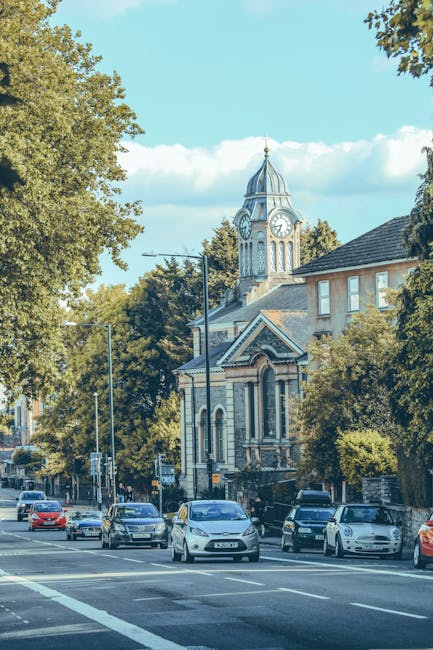 A street scene showing a row of parked cars along a pavement in a residential area, with a historic clock tower building featuring a pointed roof and decorative architectural details visible in the background. Tall trees with green foliage line the street, casting shadows over the parked vehicles and sidewalk. The sky is partly cloudy with patches of blue, and street lamps are positioned along the pavement. This setting appears to be part of a home relocation or moving process, where [COMPANY_NAME] may be involved in furniture transport or packing and loading activities, although no individuals or moving equipment are visible in this image.