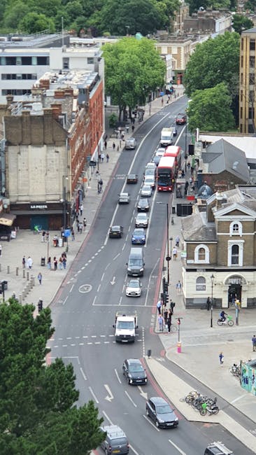 A quiet residential street in Surbiton with a gently sloping road marked with large white painted words 'KEEP CLEAR' on the asphalt. On the left side, there is a white, three-storey building with a balcony and large windows, situated close to the pavement, which has a tactile paving strip for accessibility. Further along the street, there are various terraced houses with dark tiled roofs and chimneys, some with small front yards or steps, leading up from the pavement. The street is lit by the warm glow of the setting sun, casting an orange hue on the clouds in a partly cloudy sky, enhancing the peaceful atmosphere suitable for home relocation planning. In the background, cars are parked along the roadside, and the scene suggests a typical urban environment suitable for local house removals and furniture transport operations.